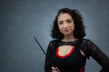 Brunette witch wearing black and red costume, pointing with magic wand, looking at camera. Ideal for Halloween web banner, trick or treat, spooky advertising. Horizontal studio portrait, copy space.