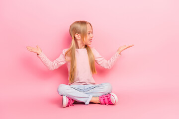 Adorable smiling girl sitting against a pink background showing cheerful gestures in casual wear and trendy sneakers