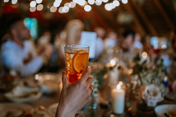 A hand holding a glass of iced cocktail with orange slices at a festive dinner party, surrounded by candlelight, bokeh lights, and blurred guests in the background.