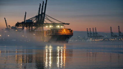 Obraz premium A cargo ship is lit up at a busy port at dawn or dusk. The harbor cranes are silhouetted against the sky, with their lights reflecting on the foggy water, creating a moody and industrial atmosphere.