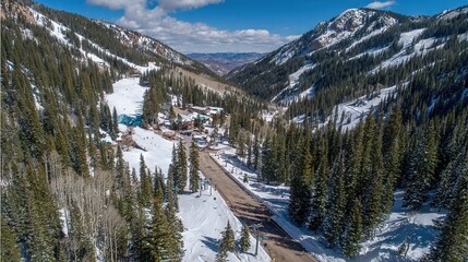 Aerial view of alpine valley with snow covered slopes, pine forests, and mountain road leading to small settlement, serene winter landscape under clear sky