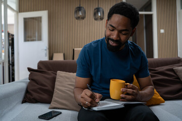 Smiling African man writing ideas while drinking coffee in living room, enjoying relaxed and inspired lifestyle