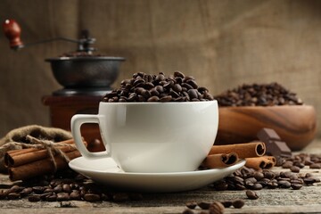 Aromatic coffee beans in cup, cinnamon and grinder on wooden table, closeup