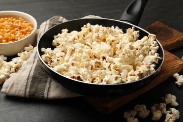 Tasty popcorn in frying pan and corn kernels on black table, closeup