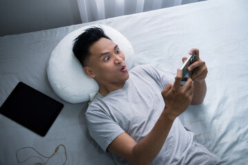 A young man lying on a bed playing games on his smartphone and shows a surprised reaction. The...
