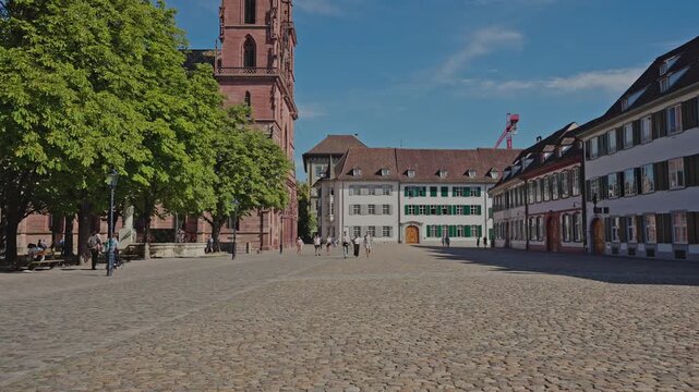 Wide view of Basel&rsquo;s M&uuml;nsterplatz with cobblestone square, historic houses, and the red sandstone Basel Minster on a sunny summer day, filmed on July 25, 2025. 