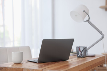 Home workspace. Laptop, cup of drink, lamp and stationery on wooden desk near window indoors