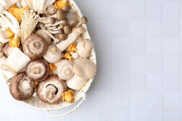 Different raw mushrooms in wicker basket on white tiled table, top view. Space for text