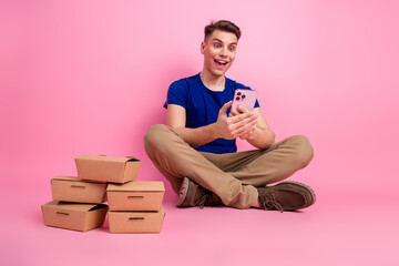 Young man in casual clothing sitting with cardboard boxes and using a phone on a bright pink...