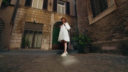 Young woman in white dress and hat with a backpack standing on cobblestone street in Rome, looking up at historic architecture with sunlight highlighting her