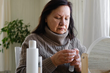 Attractive brunette senior woman putting on makeup looking in the mirror. Beautiful lady caring her face. Elderly female applied cosmetics. Natural old beauty and aging concept. Skin care and make-up