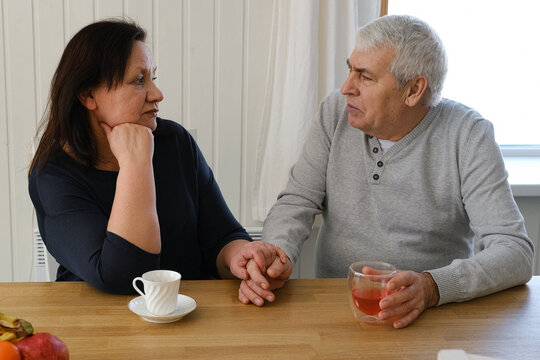 Old couple have breakfast sitting at the kitchen table, drink coffee. Embracing elderly man and woman. Senior couple together marriage in retirement talking with each other. Enjoying the pleasantries - Powered by Adobe