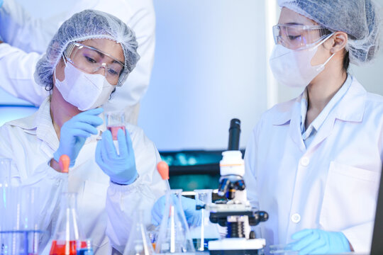 A diverse team of scientists collaborates in a genetics lab. A male researcher explains a procedure with a liquid sample while a DNA helix is displayed on a monitor behind them.
