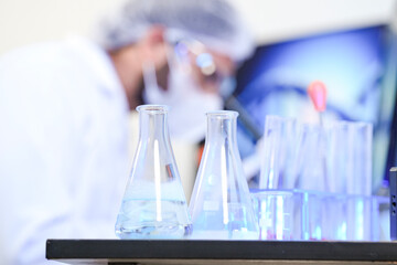 A scientist in protective gear conducts genetic research in a modern lab. He analyzes a chemical in a flask with a DNA helix displayed on a monitor in the background.
