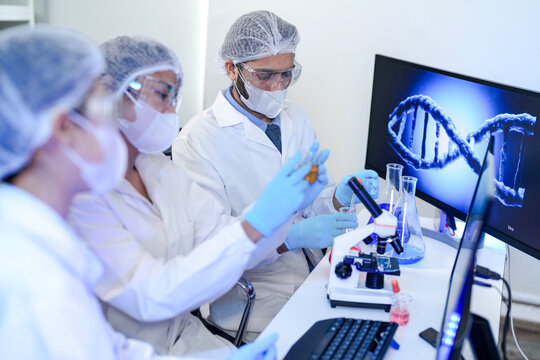 A diverse team of scientists collaborates in a genetics lab. A male researcher explains a procedure with a liquid sample while a DNA helix is displayed on a monitor behind them. - Powered by Adobe