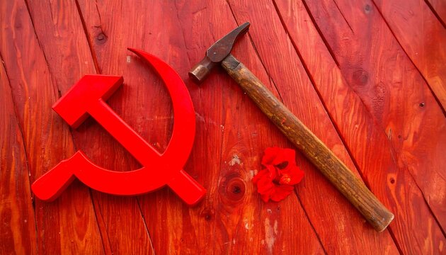 A vivid red communist symbol, the hammer and sickle, rests on a vibrant crimson wooden surface, accompanied by a weathered hammer and a delicate flower.