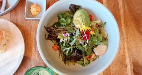 Salad bowl with avocado, mixed greens, edible flowers, cherry tomatoes on wooden table. Healthy meal, fresh produce, organic eating concept.