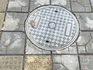 Round metal manhole cover on city sidewalk, surrounded by patterned pavement and tactile tiles, symbolizing urban infrastructure.