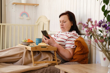 Senior woman having breakfast sitting in bed, drinking tea, enjoying her morning at home. Old lady eating from tray and using phone. Elderly female in pajamas relaxing with cup of coffee in a bedroom.