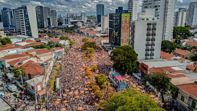 Aerial drone view of street carnival parade at Largo da Batata, Faria Lima, S&atilde;o Paulo, Brazil, with huge crowd, colorful umbrellas, festive atmosphere and city buildings captured by DJI Mavic 3