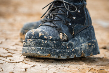 A pair of worn black platform boots covered in dried mud rests on cracked, arid ground in the...