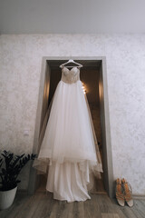 Wedding photo of long white dress hanging on hanger in room in morning with shoes.
