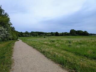 A winding gravel path leads through lush green fields and meadows under a soft, overcast sky