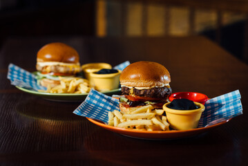 Two Oktoberfest burgers with tomato, cheese, and sauce, served with French fries and sauces. Presented on a plate with a Bavarian checkered napkin (lozenge pattern), evoking the spirit of Oktoberfest.