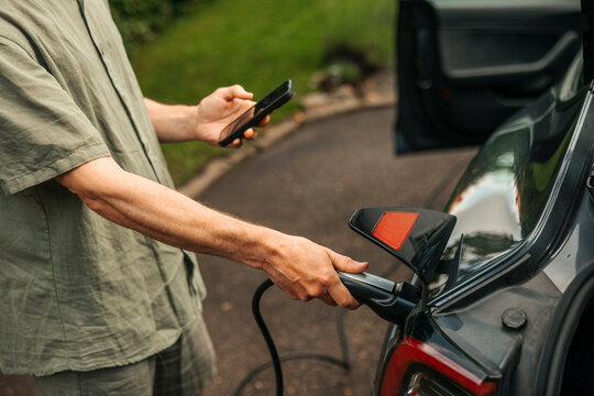 High angle view of man using smart phone while plugging in electric plug for charging electric car