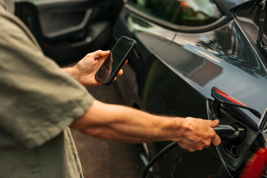 Midsection of man using mobile app while charging hybrid car with electric plug