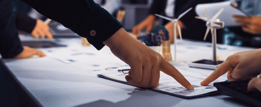 A dynamic business meeting scene showing hands pointing to documents on a table, highlighting collaboration and planning in a modern office with green energy models. SACTR