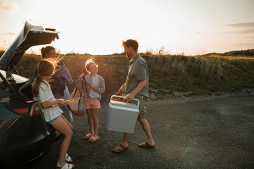 Father carrying cooler while talking with daughters standing near car at sunny day