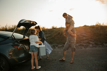 Father carrying daughter on shoulder while looking at woman unloading car trunk with girl under sunny sky