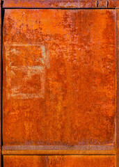 Close-up of a heavily rusted rectangular metal panel surrounded by a visible frame. The rust displays rich orange and brown tones with some lighter, faded rectangular markings on the surface.