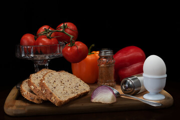 Still life composition with fresh tomatoes, whole grain bread, onion slice, colorful peppers, boiled egg in holder, spices and crystal bowl, arranged on wooden board with dark background.