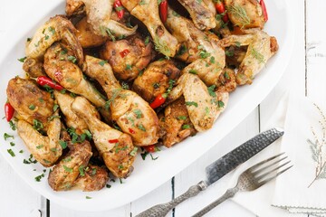 A feast of garlic herb roasted chicken pieces are arranged on a white platter. Red chili peppers add a splash of color, while antique silverware lies beside the platter on a white wooden surface