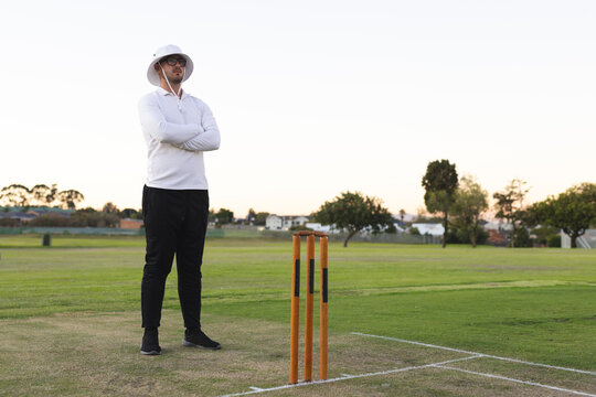 Male umpire standing near cricket stumps on pitch with crease lines in sun hat and sunglasses
