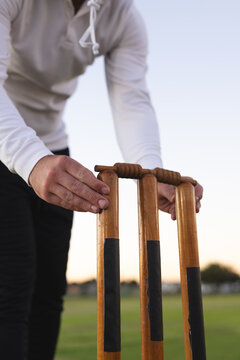 Man bending forward adjusting cricket stumps and bails on grassy sports field at dusk