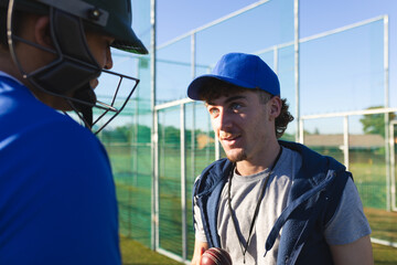 Male coach with cricket ball and guiding male player listening at practice net on grass field