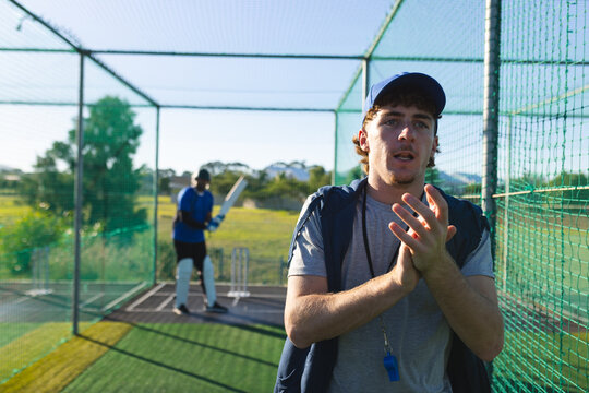 Male coach clapping, speaking toward batsman with cricket bat next to wicket in net cage