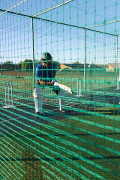 Man standing in green practice net cage in helmet and pads gripping cricket bat at stumps