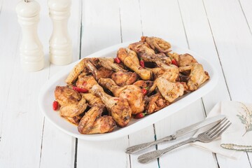 A delicious platter showcases roasted chicken pieces, seasoned and garnished with red chili peppers, presented with cutlery and spice shakers on a white wooden table