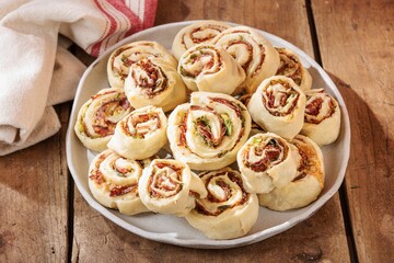 A collection of golden pinwheel pastries rests on a light gray plate. Salami and greens are rolled into the dough, creating a delicious appetizer on a rustic wooden surface