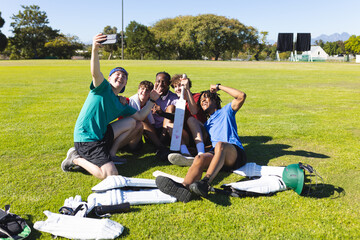 Diverse male teammates in jerseys taking selfie on sports field with smartphone and cricket bat
