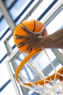 Athlete forearm gripping basketball above metal rim, net in gym under beams capturing tension