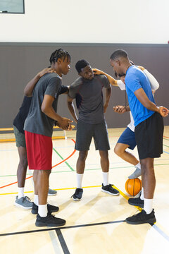 Diverse male partners huddling on gym basketball court with orange basketball and bench