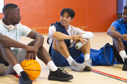Diverse male partners resting and chatting on gym court holding basketball water bottle duffel bag - Powered by Adobe