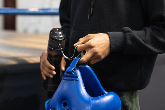 African american man in hoodie holding blue headgear and water bottle ringside in boxing gym