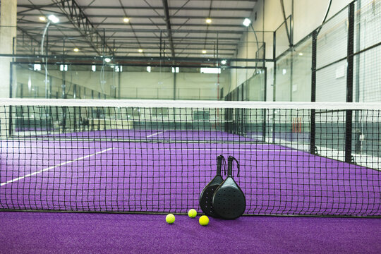Two black padel rackets are leaning by net on purple turf while three yellow balls are lying