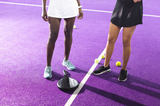 Diverse female duo standing on purple court in sport dresses next to black paddle and yellow balls